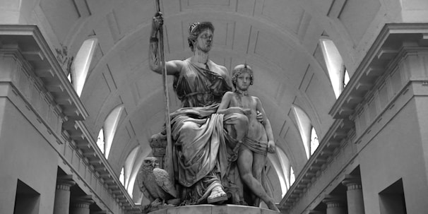 A classical sculpture of two figures in a grand architectural setting. The main figure is seated, holding a staff, with an owl at their feet, symbolizing wisdom. The other figure stands close, draped in a robe, under an arched ceiling.