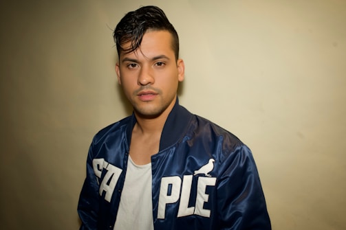 A young man with dark hair is wearing a dark blue jacket with the word 'STAPLE' written in bold white letters and a small bird logo. Underneath, he is wearing a white t-shirt. The background is a plain off-white color.