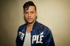 A young man with dark hair is wearing a dark blue jacket with the word 'STAPLE' written in bold white letters and a small bird logo. Underneath, he is wearing a white t-shirt. The background is a plain off-white color.