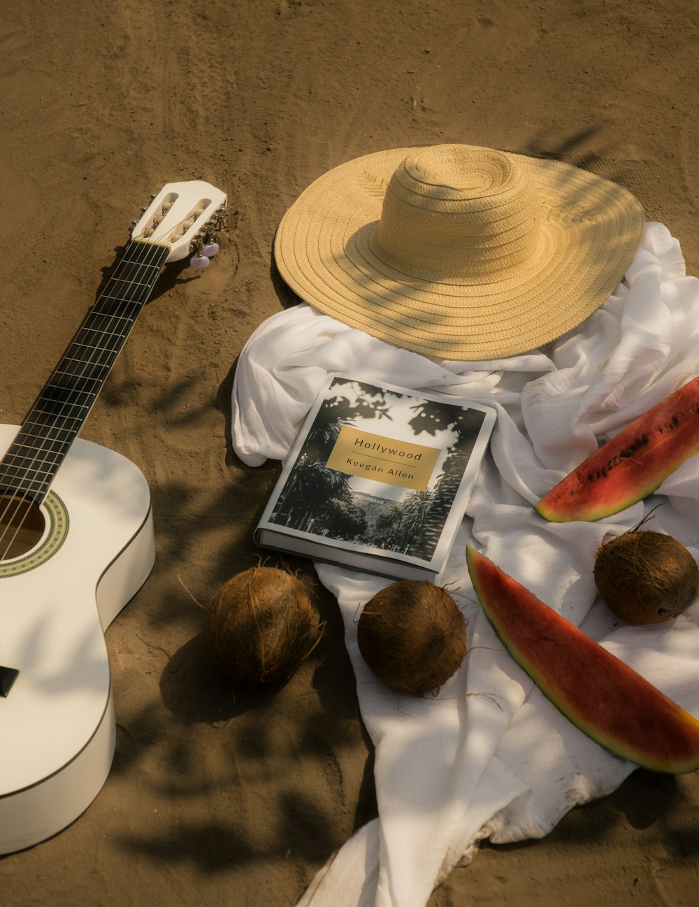 a guitar, hat, watermelon, and a book on a beach