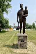 A bronze statue of a man wearing a fez hat and holding a child is centered on a grassy area. The man holds a staff in his hand. The statue is mounted on a stone pedestal with an inscription. There are trees and a few parked cars in the background, as well as a building with a prominent sign.