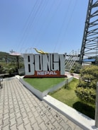 A large sign with the word 'BUNGY' in bold capital letters, with 'NEPAL' written underneath it in smaller letters. The sign is positioned on a raised landscaped area with artificial grass and surrounded by trees and shrubs. A yellow sculpture of a person in a diving pose is positioned on top of the sign. In the background, there are metal structures and wires suggesting an adventure or theme park setting.