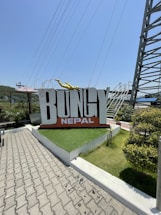 A large sign with the word 'BUNGY' in bold capital letters, with 'NEPAL' written underneath it in smaller letters. The sign is positioned on a raised landscaped area with artificial grass and surrounded by trees and shrubs. A yellow sculpture of a person in a diving pose is positioned on top of the sign. In the background, there are metal structures and wires suggesting an adventure or theme park setting.