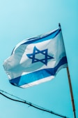 A flag featuring a blue Star of David on a white background with two horizontal blue stripes is waving against a clear blue sky. The flag is mounted on a wooden pole and gently unfurled by the breeze.