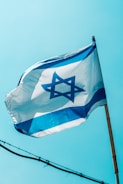 A flag featuring a blue Star of David on a white background with two horizontal blue stripes is waving against a clear blue sky. The flag is mounted on a wooden pole and gently unfurled by the breeze.