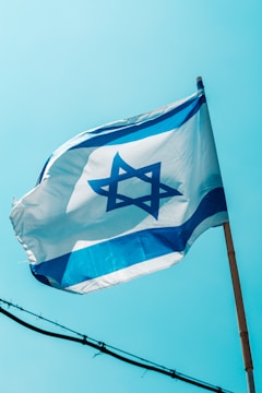 A flag featuring a blue Star of David on a white background with two horizontal blue stripes is waving against a clear blue sky. The flag is mounted on a wooden pole and gently unfurled by the breeze.