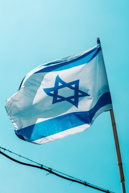 A flag featuring a blue Star of David on a white background with two horizontal blue stripes is waving against a clear blue sky. The flag is mounted on a wooden pole and gently unfurled by the breeze.