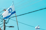 A flag with a Star of David and blue stripes on a white background waves in the breeze. In the background, electrical poles and wires are visible against a clear blue sky. Another smaller flag can also be seen partially in the lower right corner.