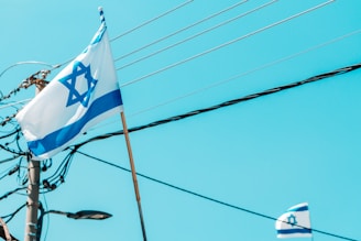 A flag with a Star of David and blue stripes on a white background waves in the breeze. In the background, electrical poles and wires are visible against a clear blue sky. Another smaller flag can also be seen partially in the lower right corner.
