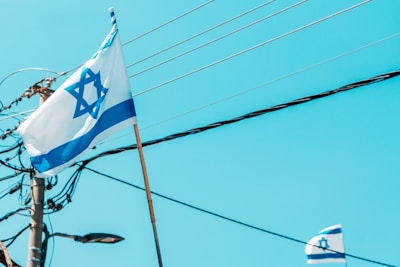 A flag with a Star of David and blue stripes on a white background waves in the breeze. In the background, electrical poles and wires are visible against a clear blue sky. Another smaller flag can also be seen partially in the lower right corner.