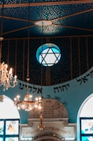 An ornate interior featuring a blue ceiling with a gold star pattern. A large chandelier hangs prominently alongside a globe-shaped lamp with a star design. Below, decorative elements include writing in Hebrew, stained glass windows, and an architectural structure with engraved symbols.