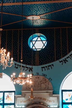 An ornate interior featuring a blue ceiling with a gold star pattern. A large chandelier hangs prominently alongside a globe-shaped lamp with a star design. Below, decorative elements include writing in Hebrew, stained glass windows, and an architectural structure with engraved symbols.