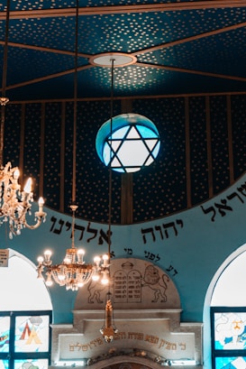 An ornate interior featuring a blue ceiling with a gold star pattern. A large chandelier hangs prominently alongside a globe-shaped lamp with a star design. Below, decorative elements include writing in Hebrew, stained glass windows, and an architectural structure with engraved symbols.