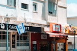 An urban scene featuring a row of small businesses along a street. A gelato shop with a black and white sign can be seen, featuring a large window front. Next door, there is a shop with a red awning and signage in a different language. Two flags, the Israeli and American, are prominently displayed, hanging from poles. The buildings are of light-colored concrete, and there is a vintage-style street lamp in the foreground.