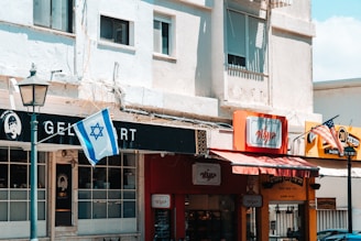 An urban scene featuring a row of small businesses along a street. A gelato shop with a black and white sign can be seen, featuring a large window front. Next door, there is a shop with a red awning and signage in a different language. Two flags, the Israeli and American, are prominently displayed, hanging from poles. The buildings are of light-colored concrete, and there is a vintage-style street lamp in the foreground.