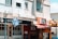 An urban scene featuring a row of small businesses along a street. A gelato shop with a black and white sign can be seen, featuring a large window front. Next door, there is a shop with a red awning and signage in a different language. Two flags, the Israeli and American, are prominently displayed, hanging from poles. The buildings are of light-colored concrete, and there is a vintage-style street lamp in the foreground.