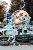A large metallic sculpture resembling a globe is adorned with multiple Israeli flags. The base of the sculpture is surrounded by dark rocks. In the background, several cars are parked, and there are trees and a blue sign with Hebrew writing.
