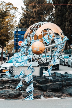 A large metallic sculpture resembling a globe is adorned with multiple Israeli flags. The base of the sculpture is surrounded by dark rocks. In the background, several cars are parked, and there are trees and a blue sign with Hebrew writing.