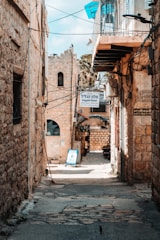 A narrow stone alleyway surrounded by aged buildings, displaying a mix of textures and colors. Signs with Hebrew text indicate local establishments, including a hotel sign. Overhead wires crisscross above, adding to the old-world charm. The alley opens up to a sunlit area with palm trees, suggesting a Mediterranean setting.