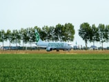 A commercial airplane is taxiing or parked on a green airfield surrounded by a row of tall trees. The aircraft has white and green livery and belongs to Transavia. In the background, there are some buildings and a partly cloudy sky.
