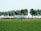 A commercial airplane is taxiing or parked on a green airfield surrounded by a row of tall trees. The aircraft has white and green livery and belongs to Transavia. In the background, there are some buildings and a partly cloudy sky.