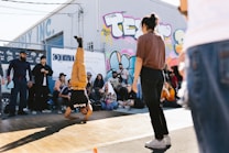 A breakdancer performs a headstand on a wooden floor outdoors, surrounded by a group of onlookers. The setting is urban, with colorful graffiti art on the wall behind them. People in casual clothing are watching, some seated, others standing. The sun casts shadows on the ground.