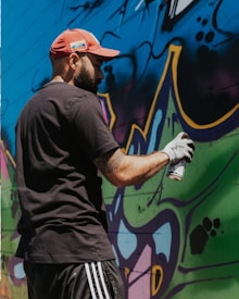 A person wearing a red cap and black shirt is spray painting a colorful mural on a wall. The mural has bold, abstract shapes with a mix of bright colors like blue, green, and purple. The person is holding a spray can in their gloved hand.