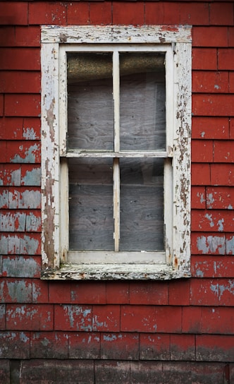 An old, weathered window framed with peeling white paint rests in a wall of red, worn wooden shingles. The texture of the wall and window frame exhibits significant wear, indicative of age and exposure to the elements.