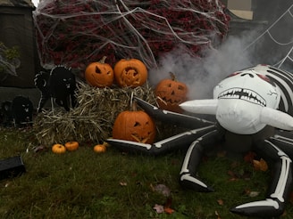 A spooky Halloween scene with several carved pumpkins placed on straw bales, surrounded by small decorative pumpkins on a grassy lawn. An inflatable spider with a skeletal pattern is positioned menacingly beside the pumpkins. Black cat silhouettes and cobwebs add to the eerie atmosphere, with artificial fog providing a misty backdrop.