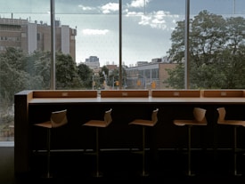 A row of modern wooden stools is lined up against a counter facing large windows. Outside, trees and urban buildings are visible under a slightly overcast sky. The area appears to be a relaxed and quiet indoor space, possibly a cafe or library, with a view of the cityscape.