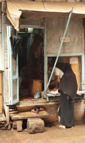 A woman in a black garment is standing in front of a small shop with a blue wooden frame. Inside the shop, a man in a white shirt is preparing meat. The shop appears rustic with a partially rolled-up canvas cover and some worn-out materials. There are bags hanging inside, and a small wooden stool is visible. The ground outside is wet, with a stone and a pair of shoes on a step in front of the shop.