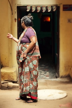 An elderly woman is standing outside a door, wearing a traditional patterned sari with floral designs. Her hair is tied in a bun, and she is looking away from the camera. The entrance is decorated with a string of traditional banners. The interior beyond the doorway is dimly lit.
