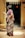 An elderly woman is standing outside a door, wearing a traditional patterned sari with floral designs. Her hair is tied in a bun, and she is looking away from the camera. The entrance is decorated with a string of traditional banners. The interior beyond the doorway is dimly lit.