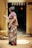 An elderly woman is standing outside a door, wearing a traditional patterned sari with floral designs. Her hair is tied in a bun, and she is looking away from the camera. The entrance is decorated with a string of traditional banners. The interior beyond the doorway is dimly lit.