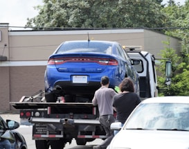 A blue car is being towed by a tow truck in a parking lot. Two people are standing nearby, possibly discussing or overseeing the towing process. The scene is set in daylight with a building and trees in the background.
