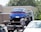 A blue car is being towed by a tow truck in a parking lot. Two people are standing nearby, possibly discussing or overseeing the towing process. The scene is set in daylight with a building and trees in the background.