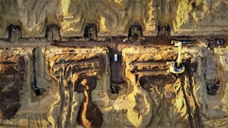 Aerial view of a large construction site with heavy machinery and vehicles. The site features multiple trenches and excavated areas with dirt and earth-moving equipment. There are visible tire marks and pathways created by the machinery. The predominance of brown and yellow hues indicates the natural earth and sand present at the location.