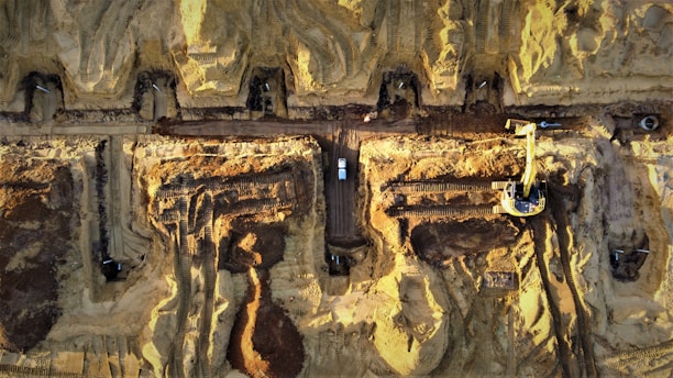 Aerial view of a large construction site with heavy machinery and vehicles. The site features multiple trenches and excavated areas with dirt and earth-moving equipment. There are visible tire marks and pathways created by the machinery. The predominance of brown and yellow hues indicates the natural earth and sand present at the location.