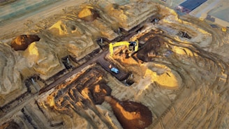 A construction site with large piles of earth and heavy machinery. An excavator with a yellow arm is actively digging beside mounds of dirt, and a small vehicle is parked nearby. The area shows clear marks of recent excavation, with tracks from machinery visible on the ground.
