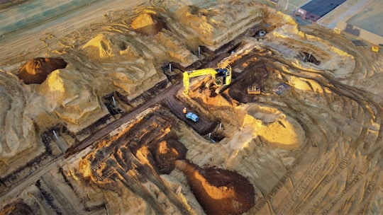 A construction site with large piles of earth and heavy machinery. An excavator with a yellow arm is actively digging beside mounds of dirt, and a small vehicle is parked nearby. The area shows clear marks of recent excavation, with tracks from machinery visible on the ground.