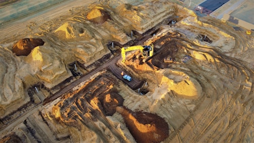 A construction site with large piles of earth and heavy machinery. An excavator with a yellow arm is actively digging beside mounds of dirt, and a small vehicle is parked nearby. The area shows clear marks of recent excavation, with tracks from machinery visible on the ground.
