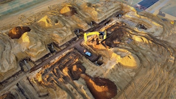 A construction site with large piles of earth and heavy machinery. An excavator with a yellow arm is actively digging beside mounds of dirt, and a small vehicle is parked nearby. The area shows clear marks of recent excavation, with tracks from machinery visible on the ground.