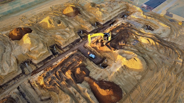 A construction site with large piles of earth and heavy machinery. An excavator with a yellow arm is actively digging beside mounds of dirt, and a small vehicle is parked nearby. The area shows clear marks of recent excavation, with tracks from machinery visible on the ground.