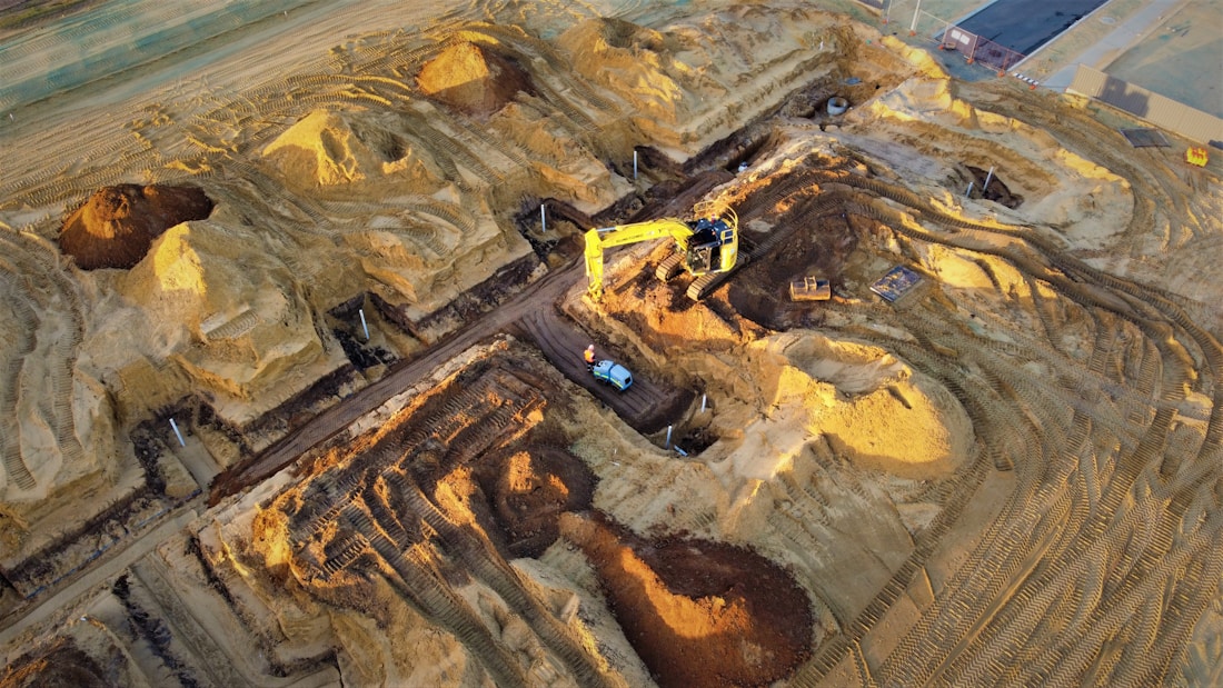 A construction site with large piles of earth and heavy machinery. An excavator with a yellow arm is actively digging beside mounds of dirt, and a small vehicle is parked nearby. The area shows clear marks of recent excavation, with tracks from machinery visible on the ground.