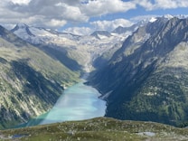 A breathtaking mountainous landscape featuring a long, winding turquoise lake nestled between towering peaks. Snow-capped mountains stand majestically in the distance under a partly cloudy blue sky. Verdant greenery covers the lower slopes, adding a lush contrast to the rugged rock formations.