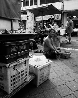 A street market scene featuring a shirtless man seated on the pavement next to some crates. Other vendors and shoppers are visible in the background, with signage in a foreign language above. An umbrella provides shade for some of the individuals.