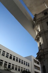 A blend of modern and classical architecture is captured, featuring a contemporary building with straight lines and rectangular windows on the left and a historic, ornate building with detailed masonry on the right. Above, a sleek, wide flat structure or bridge element contrasts with the blue sky, adding a dynamic element.