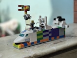 A colorful LEGO creation resembling a vehicle with a police label on the front. It features a playful mix of colors including white, blue, orange, green, and black bricks. There is a small wheel and intricate components on top and it sits on a marble surface.