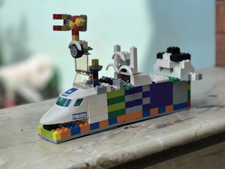 A colorful LEGO creation resembling a vehicle with a police label on the front. It features a playful mix of colors including white, blue, orange, green, and black bricks. There is a small wheel and intricate components on top and it sits on a marble surface.