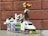 A colorful Lego model resembling a vehicle with a 'Police' label on the front. It has a mix of various bricks in colors like purple, orange, green, and blue, forming the base and body. On top, a multicolored structure with a wheel adds a playful detail. The background is a brown brick wall.